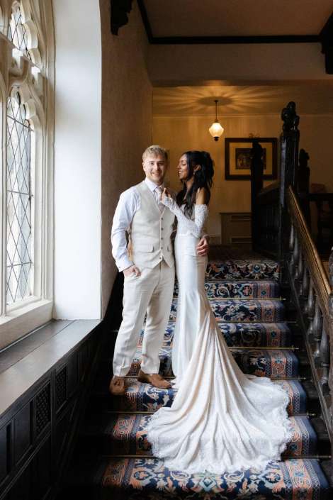bride and groom on staircase A bride in a long white wedding gown with a flowing train stands with a groom in a light-colored suit on a patterned carpeted staircase inside a historic building.