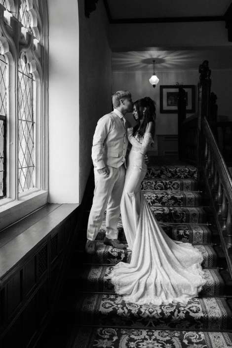 bride and groom embrace on staircase Bride in a long lace wedding gown and groom in a light suit embrace on a patterned staircase beside a large arched window.