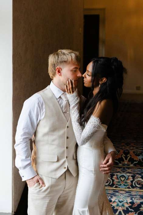intimate wedding kiss Bride in a white lace-sleeve wedding dress gently cupping the groom's face as they share a tender moment indoors.