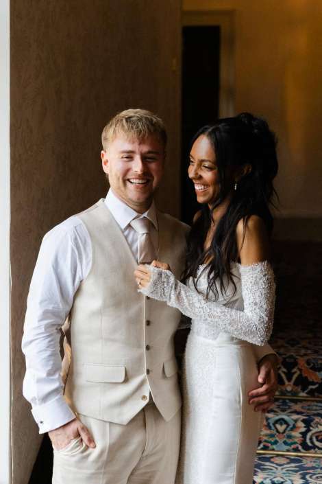 joyful bride and groom portrait A bride in a white lace wedding dress stands next to a groom in a light beige suit, both smiling indoors.
