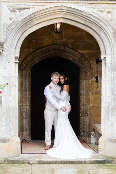 couple at stone archway A bride in a white wedding dress and groom in a beige suit embrace under a stone arch doorway.