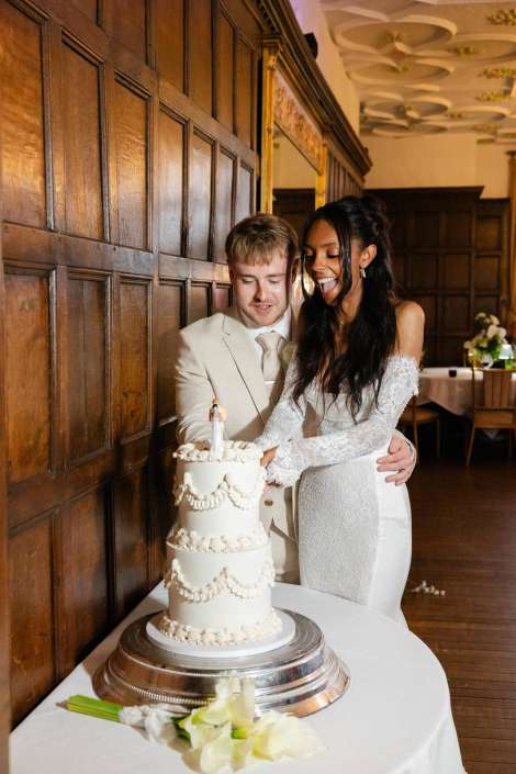 bride and groom cutting cake A bride and groom cut a tall white wedding cake together beside a wooden-paneled wall.