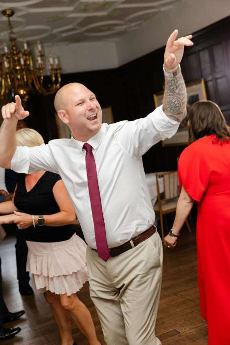 energetic dancer at reception A bald man in a white shirt and magenta tie dances with his arms raised at a party, his left arm tattoo visible.