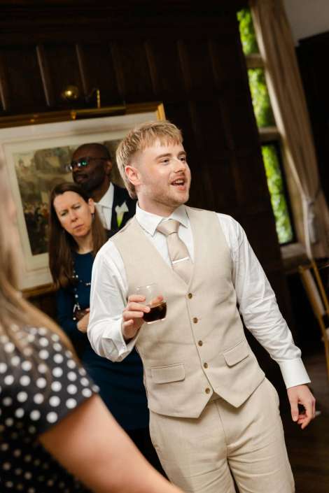 beige suit wedding toast A man in a beige suit and light tie holds a glass of wine at a formal indoor event.