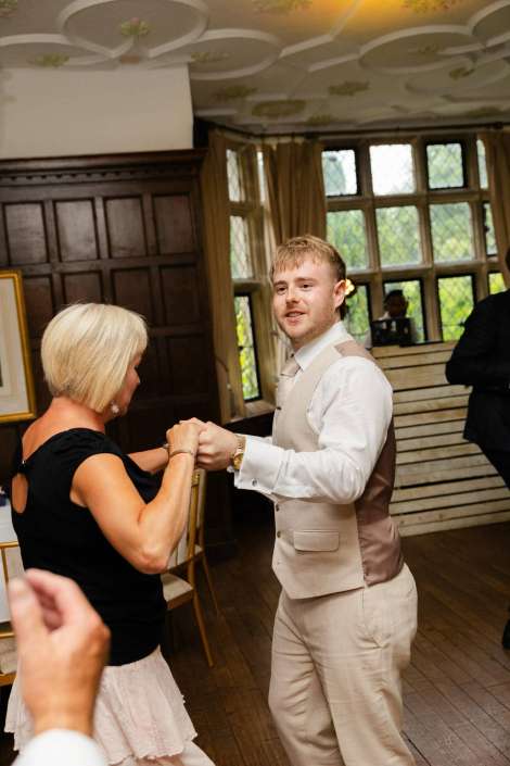 dancing couple at reception A man in a light beige suit dances with a woman in a black top, holding hands in a wood-paneled room with large windows.