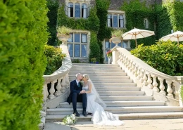 A bride in a white wedding gown and a groom in a dark suit sit together on the stone steps of an ivy-covered mansion.