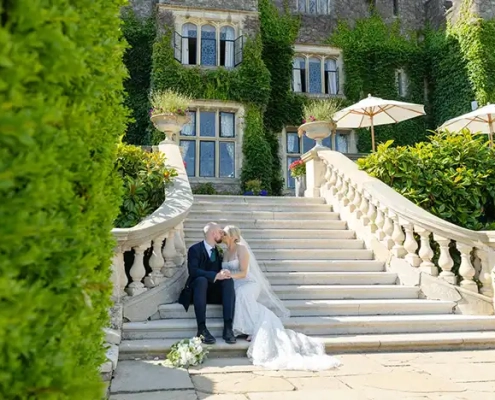 A bride in a white wedding gown and a groom in a dark suit sit together on the stone steps of an ivy-covered mansion.