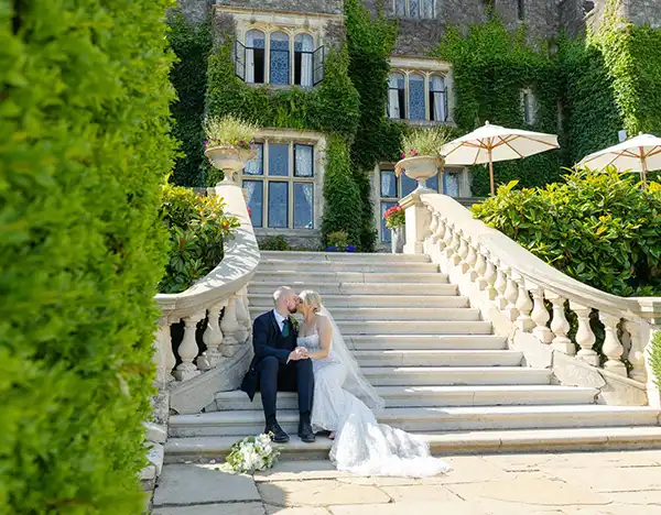 sarah & kieran, eastwell manor, louisa dettmer photography A bride in a white wedding gown and a groom in a dark suit sit together on the stone steps of an ivy-covered mansion.