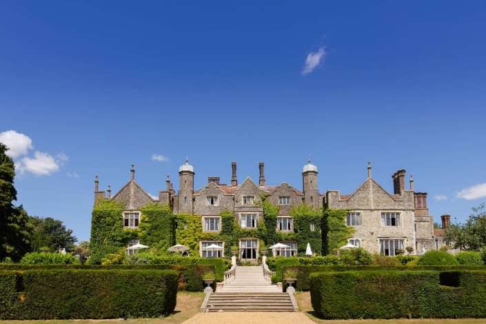 ivy covered manor house An ivy-covered historic stone mansion with a central staircase and neatly trimmed hedges beneath a bright blue sky.