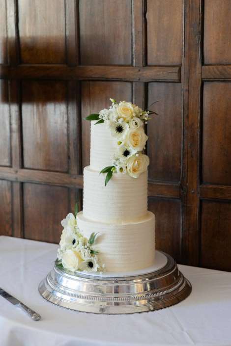 elegant white wedding cake Three-tier white wedding cake adorned with ivory roses and white blooms on a silver cake stand.