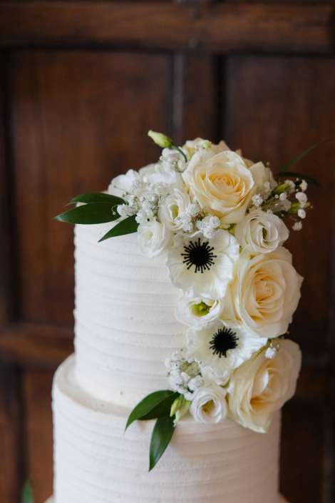 cascading white rose cake White tiered wedding cake with a cascading arrangement of cream roses, white anemones, baby’s breath, and green leaves.