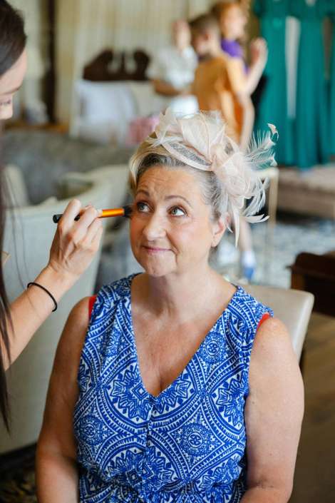 wedding day makeup session A woman in a blue patterned dress sits as a makeup artist applies eye makeup, wearing a beige feathered fascinator.