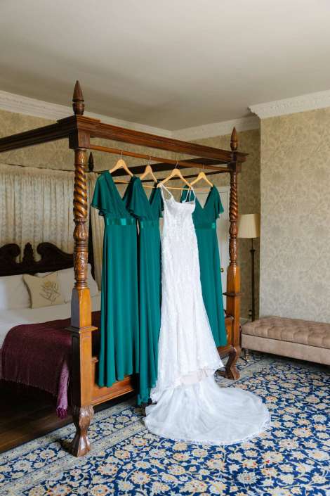 emerald bridesmaid dresses and gown A white lace wedding dress with a long train hangs beside teal bridesmaid dresses on a wooden four-poster bed in an ornate bedroom.