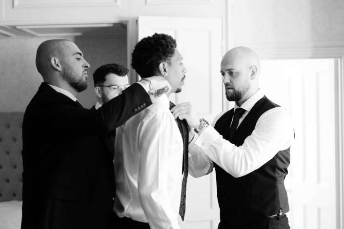 groom getting ready with groomsmen Four men in suits help adjust a tie together in a bright indoor setting.