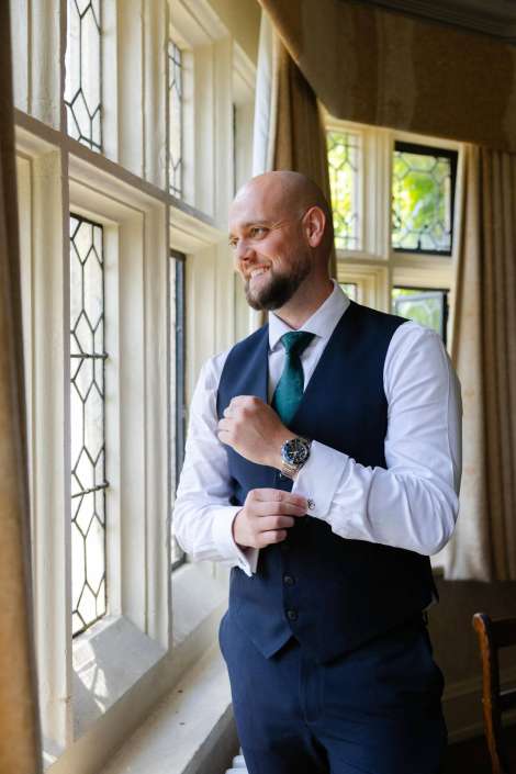 man adjusting cuff by window A bald man in a navy vest, white shirt, and teal tie stands by a sunlit window, adjusting his cuff and wearing a watch.