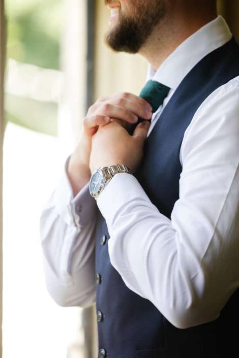 man adjusting his tie Man in a white shirt and navy vest adjusts a green tie while wearing a silver wristwatch.