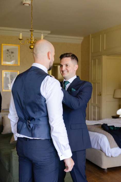 groom getting ready moment Two men in navy suits share a smile as one adjusts the other's jacket in a bedroom.