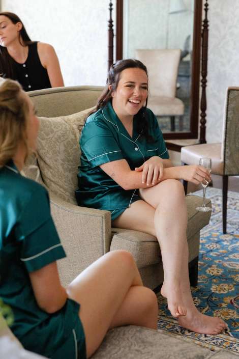 bridesmaids in satin pajamas A woman in teal satin pajamas sits on a beige sofa, smiling and holding a champagne flute.