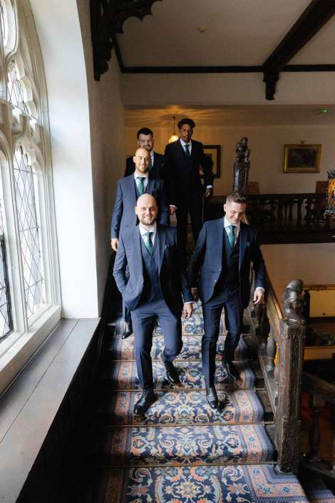 groomsmen descend grand staircase Groomsmen in navy suits and teal ties walk down a patterned carpet staircase in a historic building.
