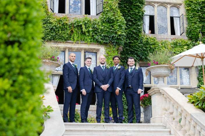 groomsmen on ivy-clad steps Five men in navy suits with green ties and boutonnieres stand together on a stone staircase in front of an ivy-covered building.