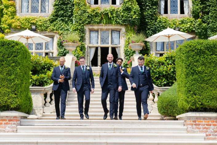wedding party descends grand staircase Five men in navy suits walk down a stone staircase in a lush ivy-covered estate courtyard.