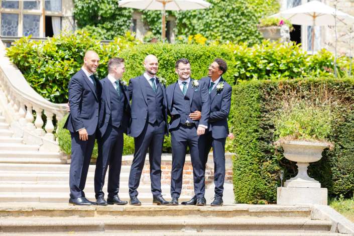groomsmen in navy suits Five men in navy suits with green ties and white boutonnieres stand together on a sunlit stone staircase in a lush garden.