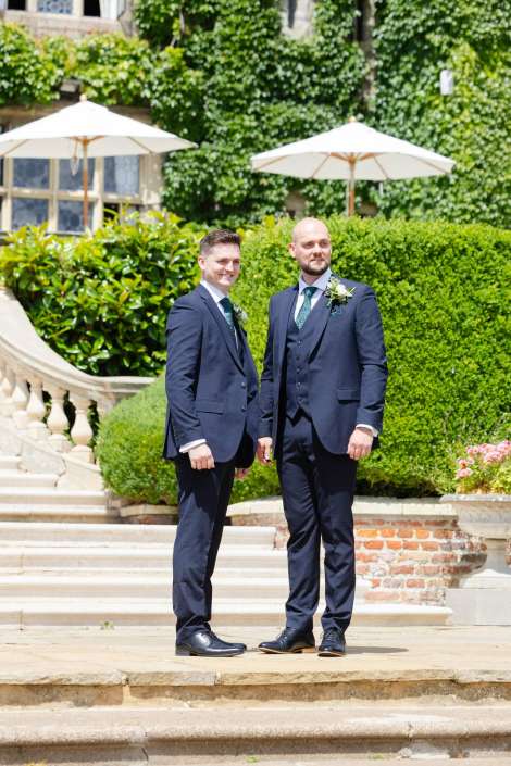 groomsmen in navy suits Two men in navy suits with boutonnieres and green ties stand on stone steps in a lush garden with hedges and white patio umbrellas.