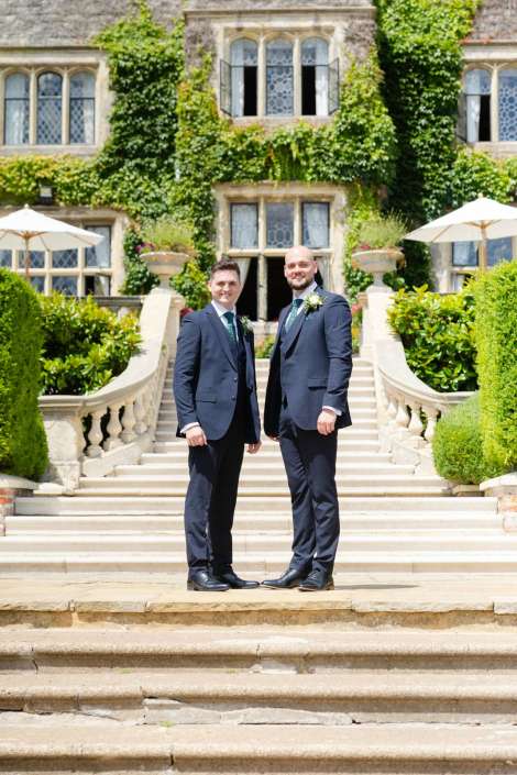 two grooms at ivy estate Two men in navy suits with boutonnières stand on stone steps in front of an ivy-covered historic building.