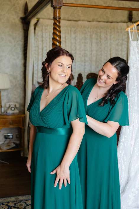 bridesmaids helping each other Two bridesmaids in teal dresses stand indoors, with one smiling as the other adjusts her gown.