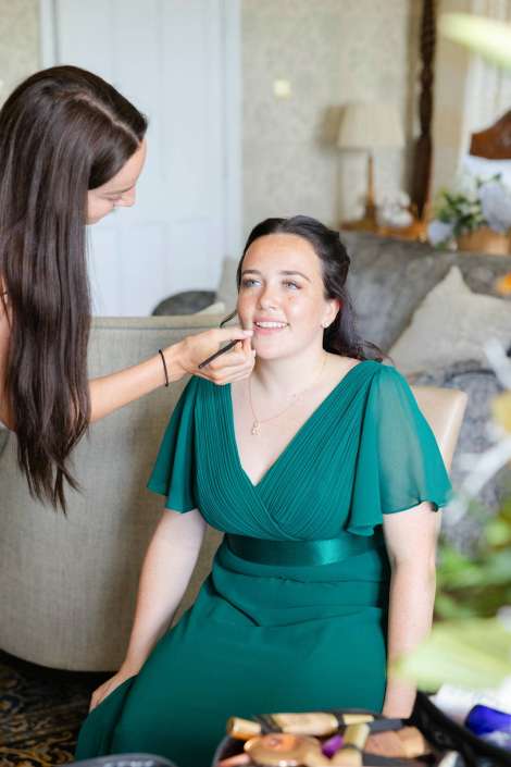 makeup artist applying lip color A woman in a teal dress sits smiling as a makeup artist applies makeup to her face.
