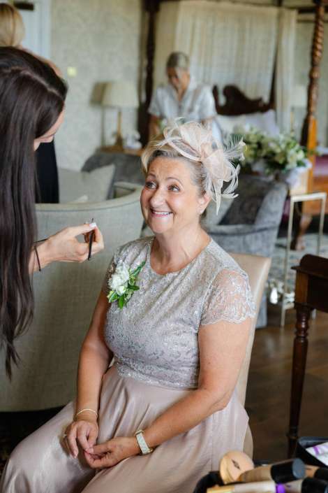 mother of the bride getting ready Smiling older woman in a silver lace dress with a pink fascinator sits as a makeup artist applies makeup.