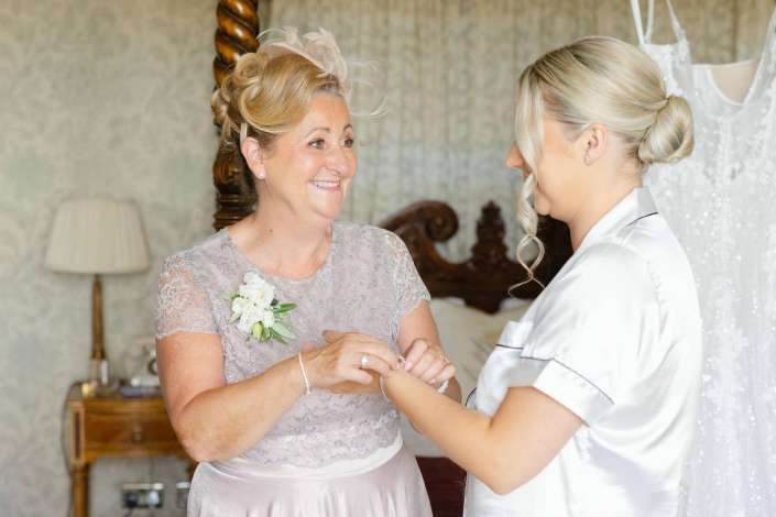 mother of the bride getting ready A smiling older woman in a pale lavender lace dress with a corsage is helped by a younger woman in a satin robe as they fasten a bracelet before the wedding.