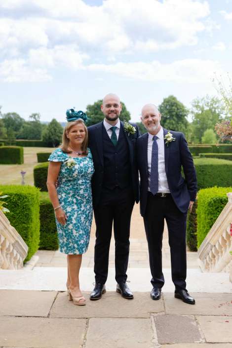 groom with parents in garden Three people in formal attire stand on an outdoor stone staircase, the woman in a teal floral dress with a teal fascinator and the two men in dark suits, with manicured hedges in the background.