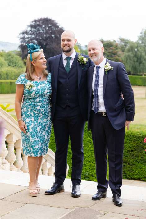 garden wedding party portrait Three people posing outdoors in formal wedding attire: a woman in teal lace dress and fascinator flanked by two men in dark suits with boutonnieres.