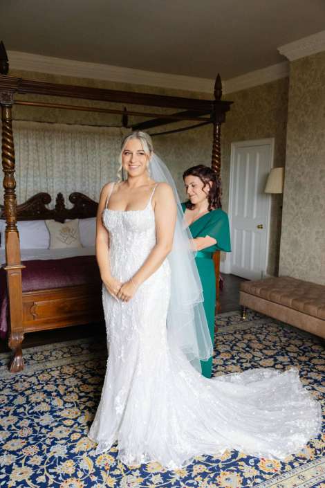 bride in white wedding gown Bride in a white lace wedding gown with veil stands in a bedroom as a bridesmaid in teal helps adjust her dress.