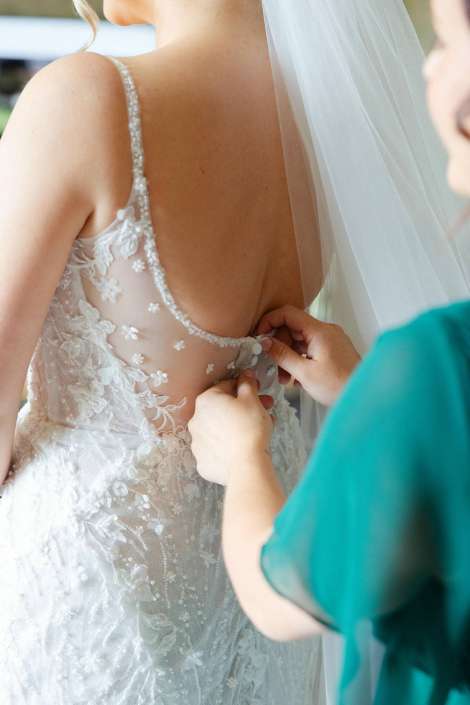 brides dress being buttoned Close-up of a bride's back as a helper fastens the floral lace wedding gown with a veil beside her.