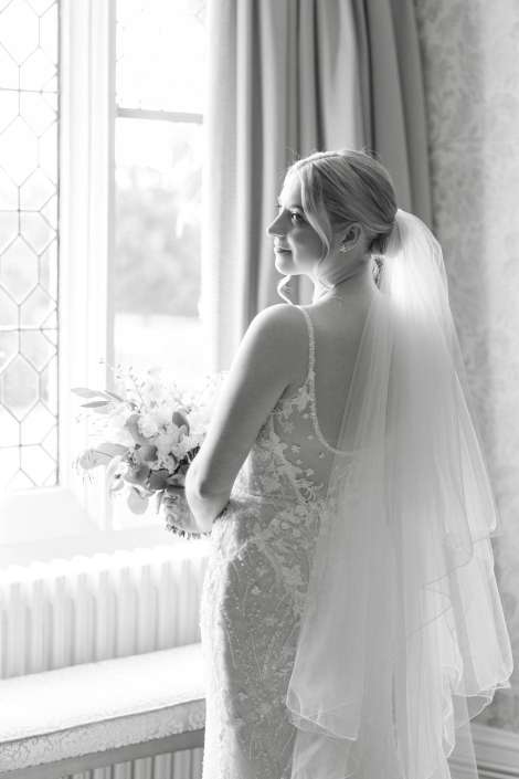 bride by window with veil Bride in a lace wedding gown with a veil stands by a window, holding a bouquet.