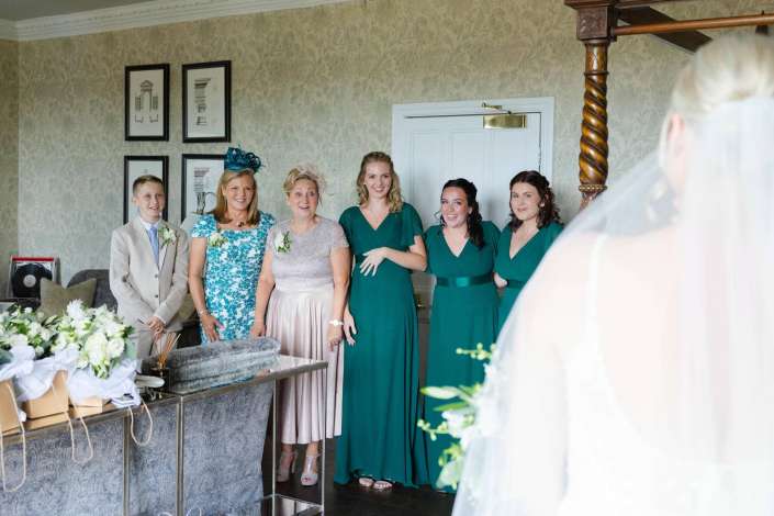 bridal party portrait Bride in a veil stands with bridesmaids in emerald green dresses and a young man in a beige suit in a vintage wallpapered room.
