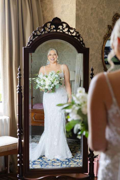 bride reflected in mirror Smiling bride in a white lace wedding dress stands in front of an ornate full-length mirror, holding a white bouquet.