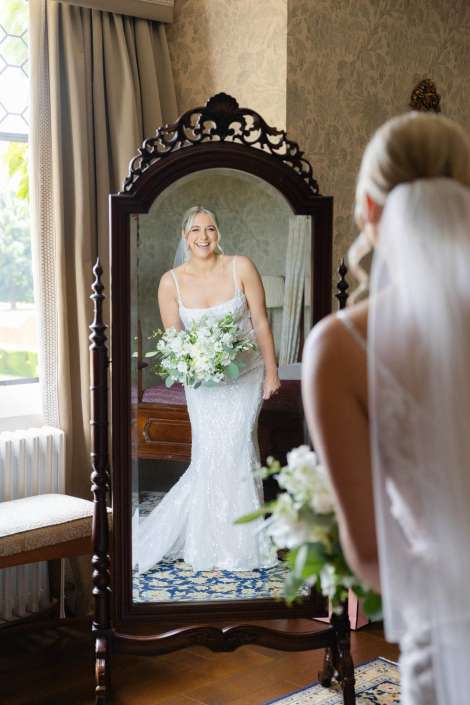 smiling bride in mirror Bride in a sparkling white wedding gown and veil smiles at her reflection in an ornate freestanding mirror while holding a white bouquet.