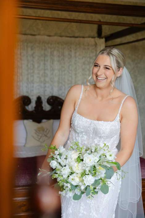 bride with white bouquet Smiling bride in a lace wedding dress with a veil, holding a white and green bouquet indoors.