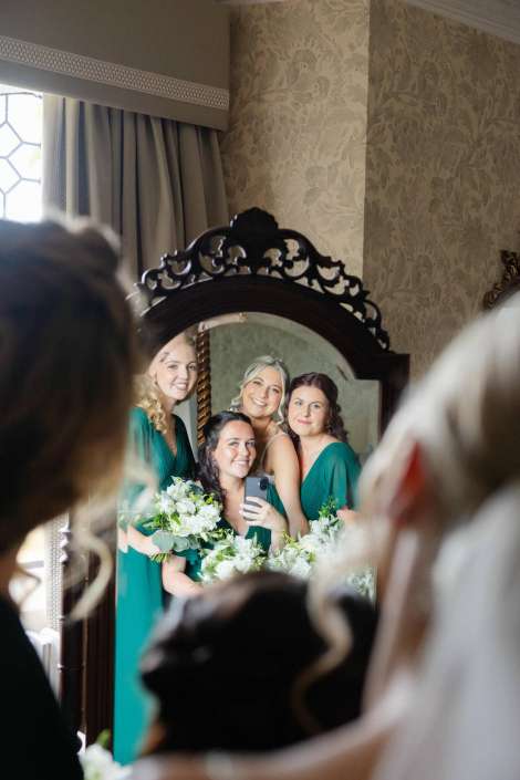 bridesmaids reflected in mirror Four bridesmaids in teal dresses smile and pose for a selfie in front of an ornate mirror, holding white bouquets.