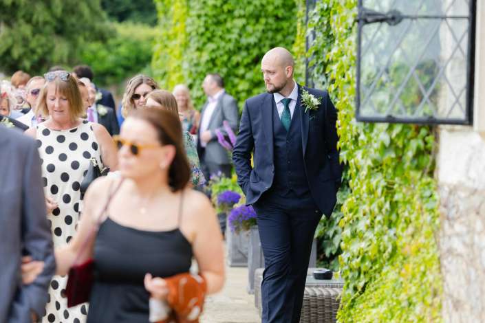 groom walking among guests A man in a navy suit with a boutonniere walks along a path beside an ivy-covered wall, while wedding guests mingle nearby.