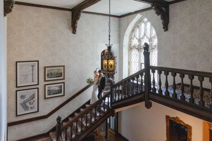 bride and groom descending staircase Bride and groom descend a dark wooden staircase in a historic room with an ornate arched window and framed artwork on patterned wallpaper.