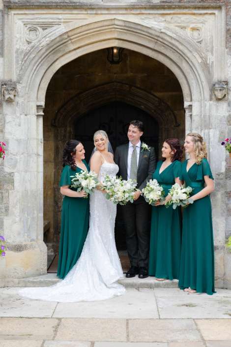 wedding party under stone arch Bride in a white lace wedding gown with a veil stands beside a groom in a dark suit, surrounded by three bridesmaids in emerald green dresses, all holding white bouquets under a carved stone arch.