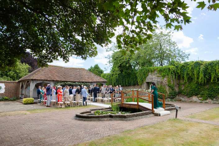 sunlit garden wedding ceremony An outdoor wedding ceremony in a brick courtyard under leafy trees, with guests seated and standing near a wooden footbridge over a small lily-filled pool.