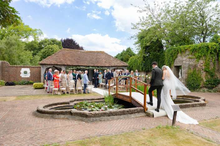 wedding ceremony on wooden bridge Bride in a white wedding gown with a long veil crosses a wooden bridge over a lily-filled pond toward guests at an outdoor wedding.