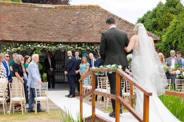 bride and groom processional Outdoor wedding ceremony with guests watching as a bride in a white gown and veil walks up a wooden aisle under a flower-decorated arch.