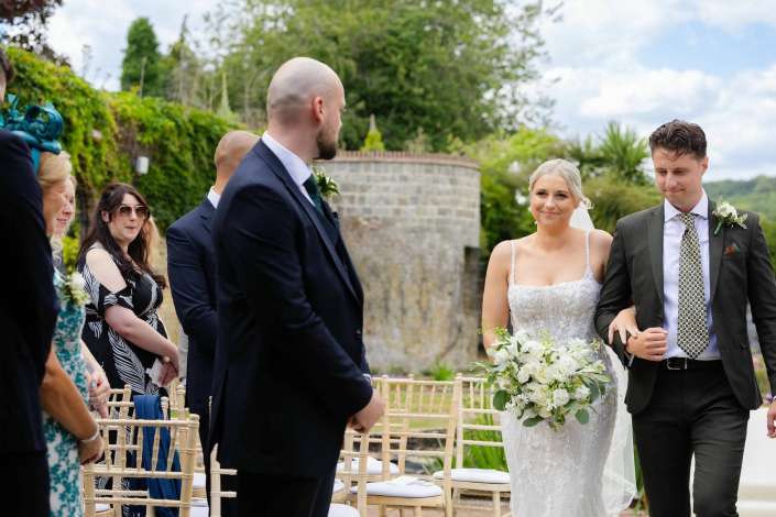 bride and groom at outdoor wedding Bride in a lace wedding dress and groom in a suit stand outdoors at their wedding, with guests seated along the aisle.