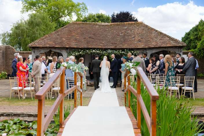 rustic outdoor wedding ceremony Bride and groom exchange vows under a rustic thatched-roof shelter as guests sit along a white aisle at an outdoor wedding.